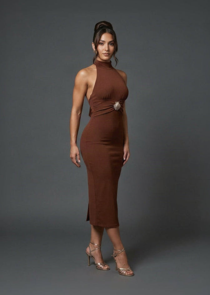 A woman modeling a fitted, sleeveless halter-neck midi dress in a rich rust brown color, featuring a decorative silver-toned circular accent at the waist. She is wearing clear high-heeled sandals against a dark gray studio backdrop.
