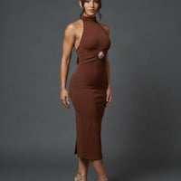 A woman modeling a fitted, sleeveless halter-neck midi dress in a rich rust brown color, featuring a decorative silver-toned circular accent at the waist. She is wearing clear high-heeled sandals against a dark gray studio backdrop.